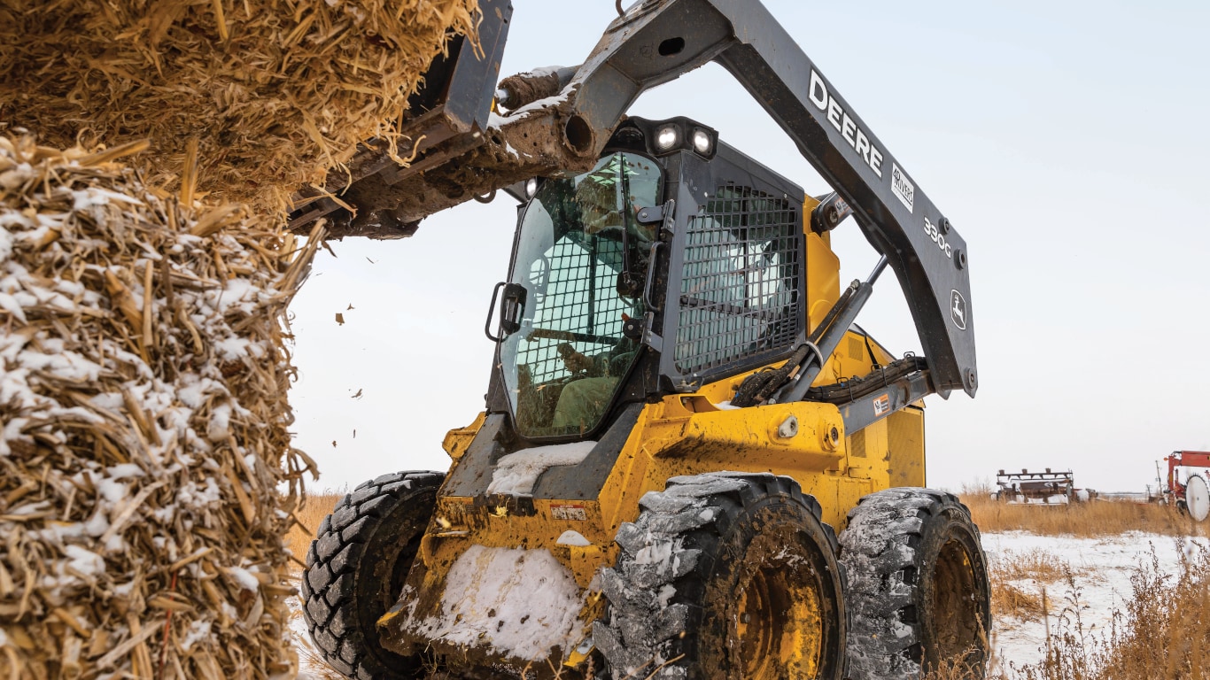 John Deere 330G Skid Steer with bale spears attachment lifting a hay bale in a snowy field. 